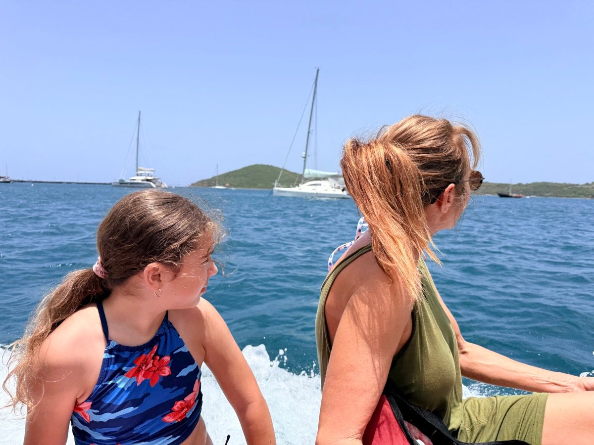 A mother and daughter looking out over the water from the back of the boat underway, sailboats and a green island beyond